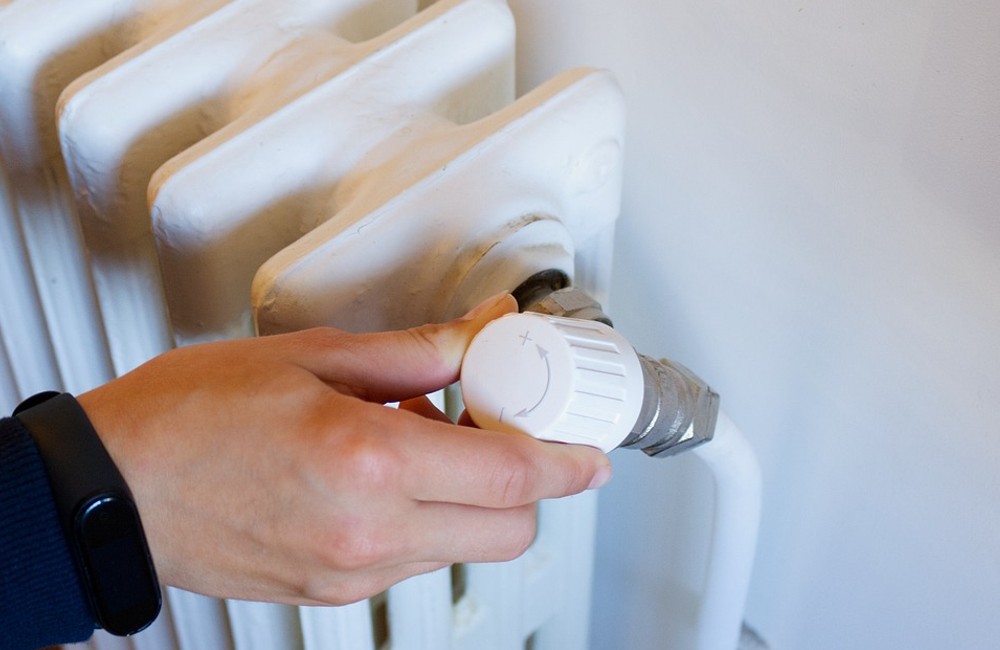 Hand dreht an einem weißen Heizkörper-Thermostat, eingestellt auf plus.